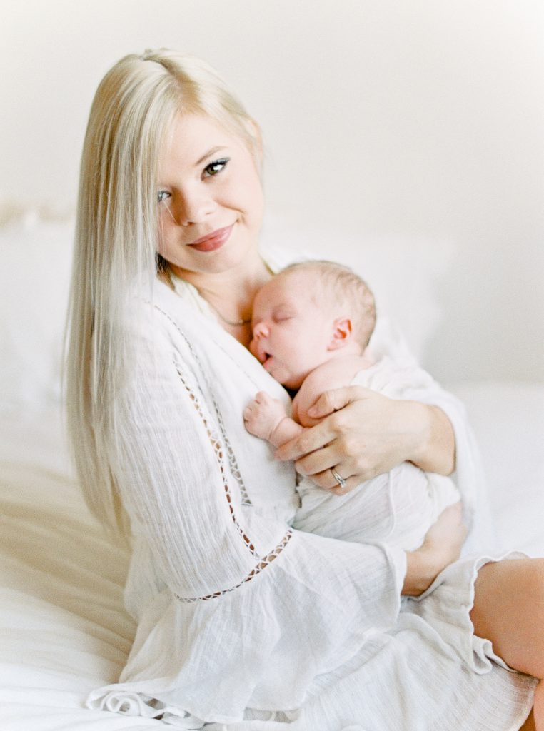 a mother with blonde hair in a white dress holds her baby in her arms as he sleeps