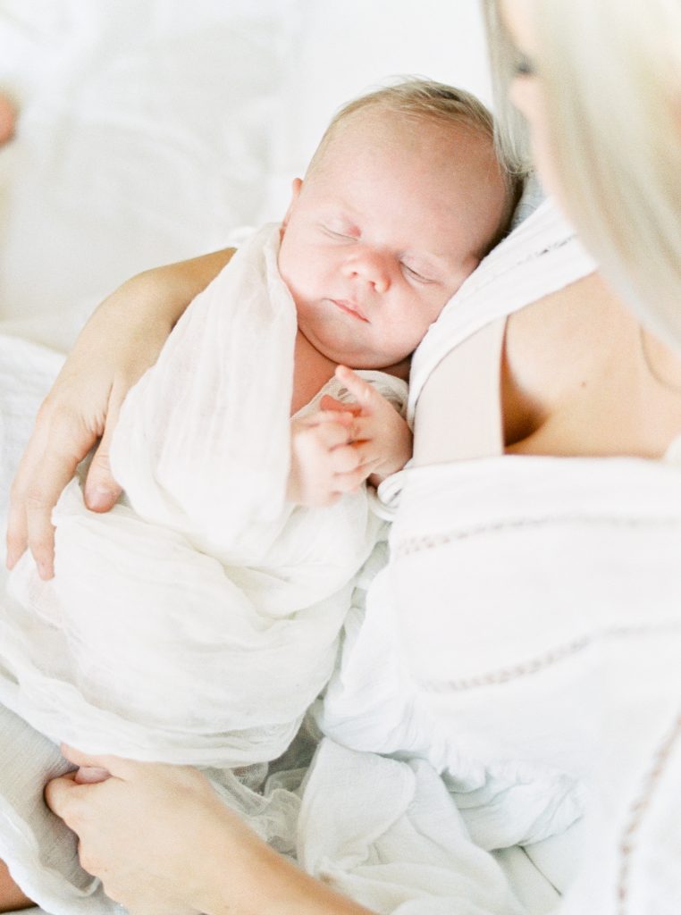 A baby sleeps in his mother's arms swaddled in a white blanket